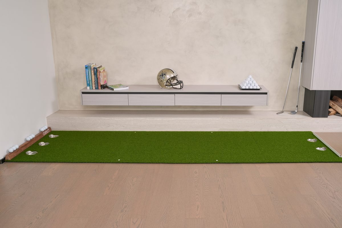 Chiputt 3 ft × 11 ft putting mat with the 2.5 ft long Dual-Hole Chipping Mat connecting to the front — total putting distance 13.5 ft, 2 holes on the chipping mat side, 3 holes on the other side, enabling two-way putting practice with the side rail as backstop on the chipping mat side; modern living room with floating credenza, books, Saints football helmet, and putters