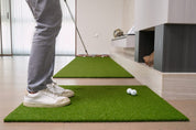 A golfer chipping from the Dual-Hole Chipping Mat onto the Chiputt putting mat — the 2 holes on the chipping mat are hidden with turf inserts for chipping practice, set in a modern living room with fireplace