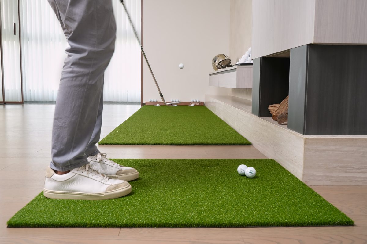 A golfer chipping from the Dual-Hole Chipping Mat onto the Chiputt putting mat — the 2 holes on the chipping mat are hidden with turf inserts for chipping practice, set in a modern living room with fireplace