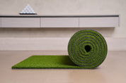 Chiputt tour-grade golf putting mat rolled up on a light wood floor beside its unrolled edge, showing how the mat rolls away for easy storage in a modern home practice setup, with a pyramid of golf balls on the credenza behind