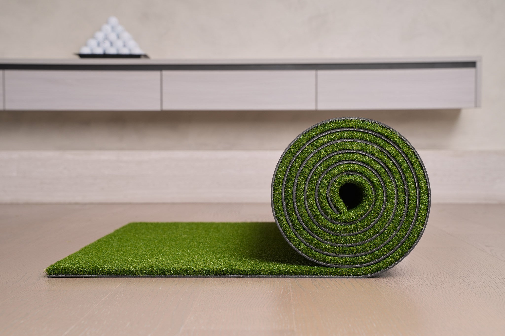 Chiputt tour-grade golf putting mat rolled up on a light wood floor beside its unrolled edge, showing how the mat rolls away for easy storage in a modern home practice setup, with a pyramid of golf balls on the credenza behind