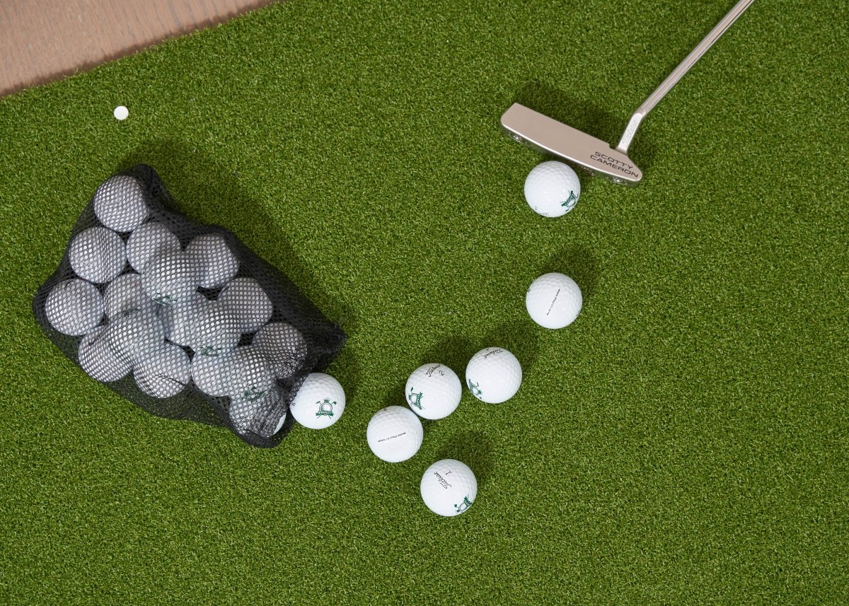 Overhead view of a Scotty Cameron putter next to several Chiputt-branded golf balls on a Chiputt mat. A mesh bag filled with additional practice golf balls lies nearby, ready for an indoor putting session.